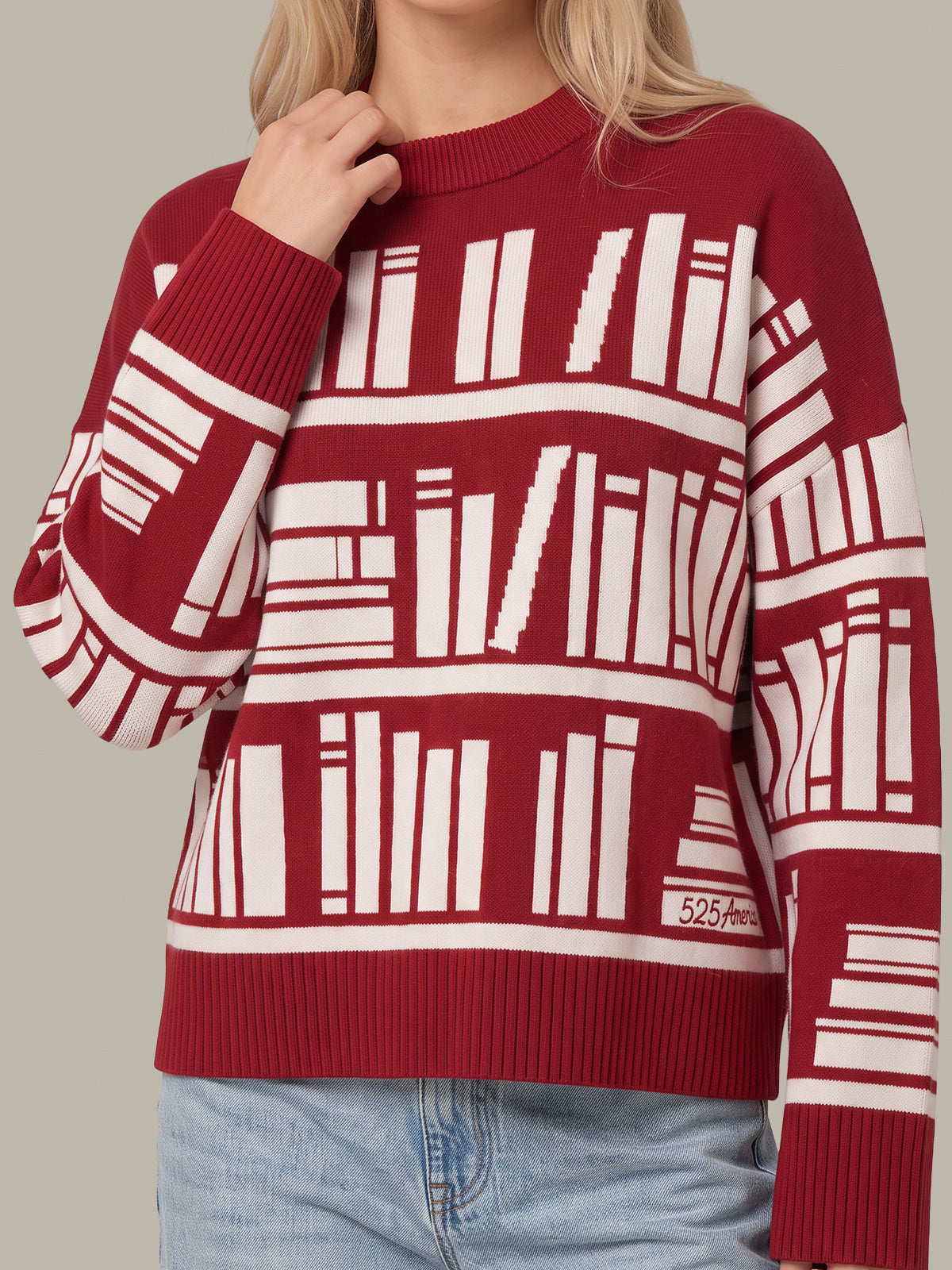 A woman wearing a burgundy crewneck sweater with a white bookshelf pattern, paired with light blue jeans, shown against a solid neutral background.