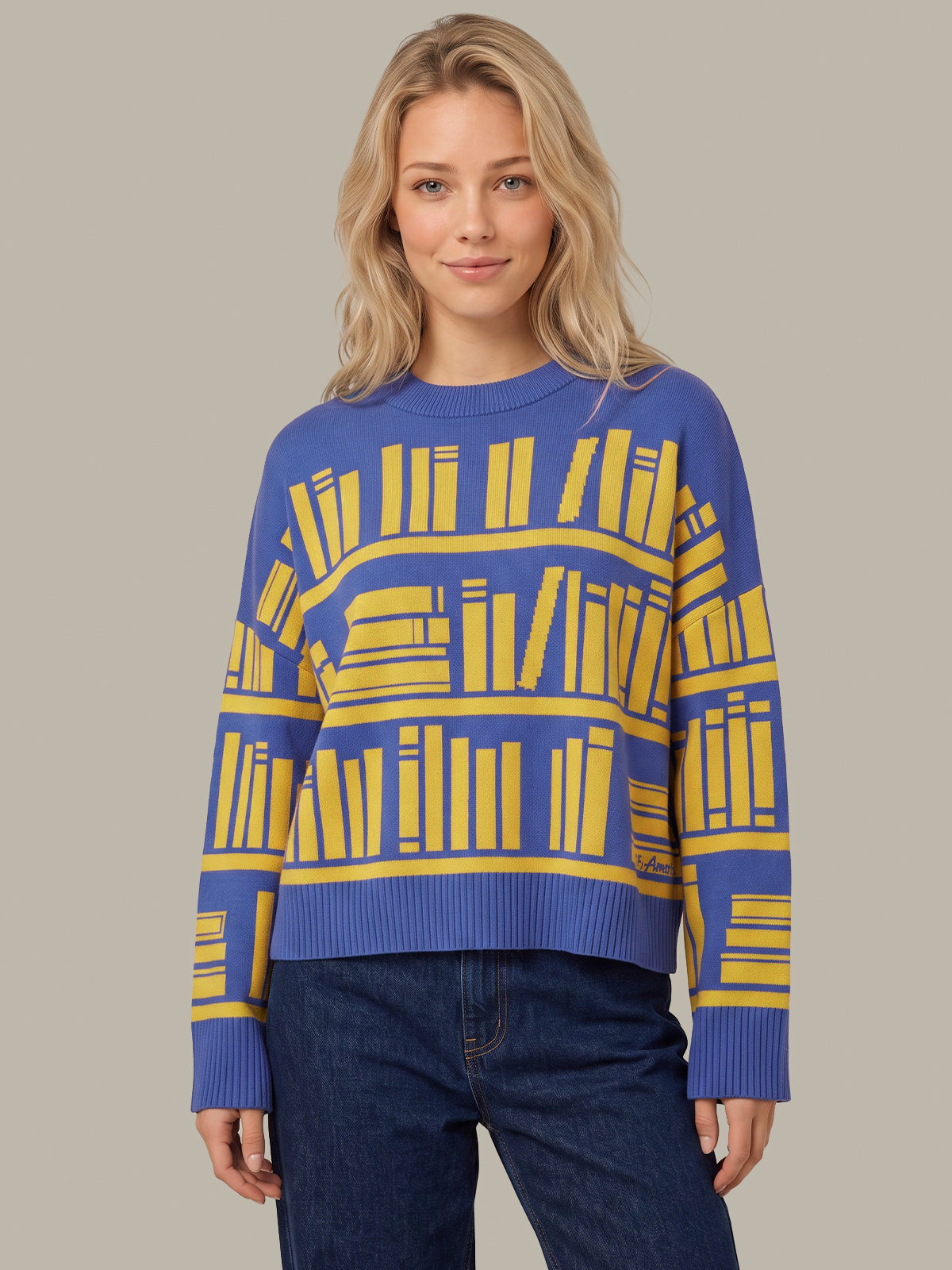A woman modeling a blue crewneck sweater with a yellow graphic pattern of book spines, paired with dark blue jeans, against a solid light gray background.