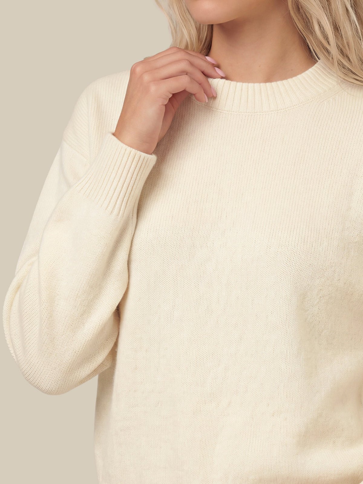 A close-up of a woman wearing a cream-colored knit crewneck sweater, showing the ribbed collar, long sleeves, and soft texture against a neutral beige background.