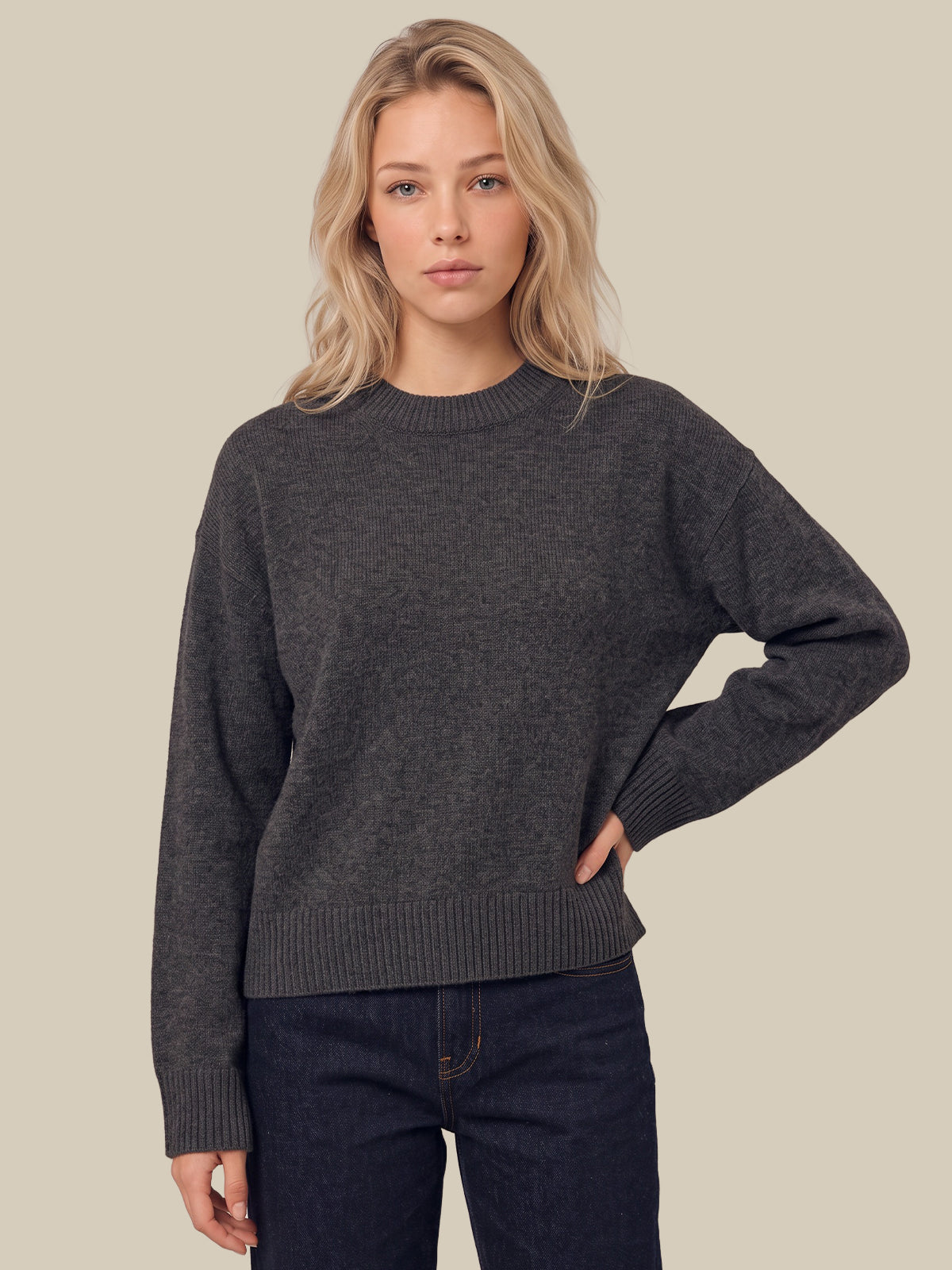 A woman modeling a dark charcoal gray crewneck sweater with ribbed cuffs and hem, paired with dark blue jeans, against a solid beige background.