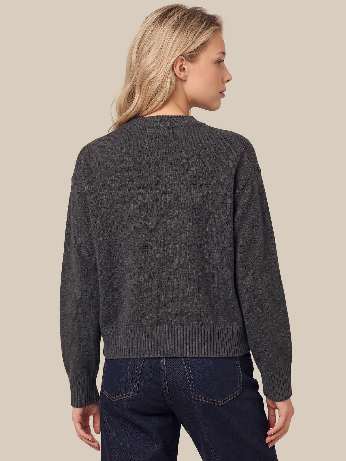 A woman shown from the back wearing a dark charcoal gray knit sweater and dark blue jeans, set against a solid light beige background.