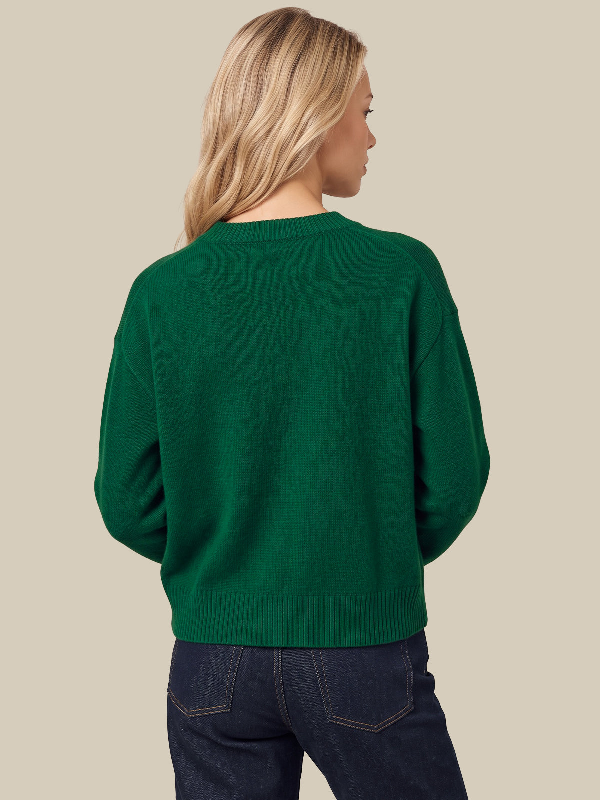 A woman shown from the back wearing a dark green crewneck sweater and dark blue jeans, set against a solid light beige background.