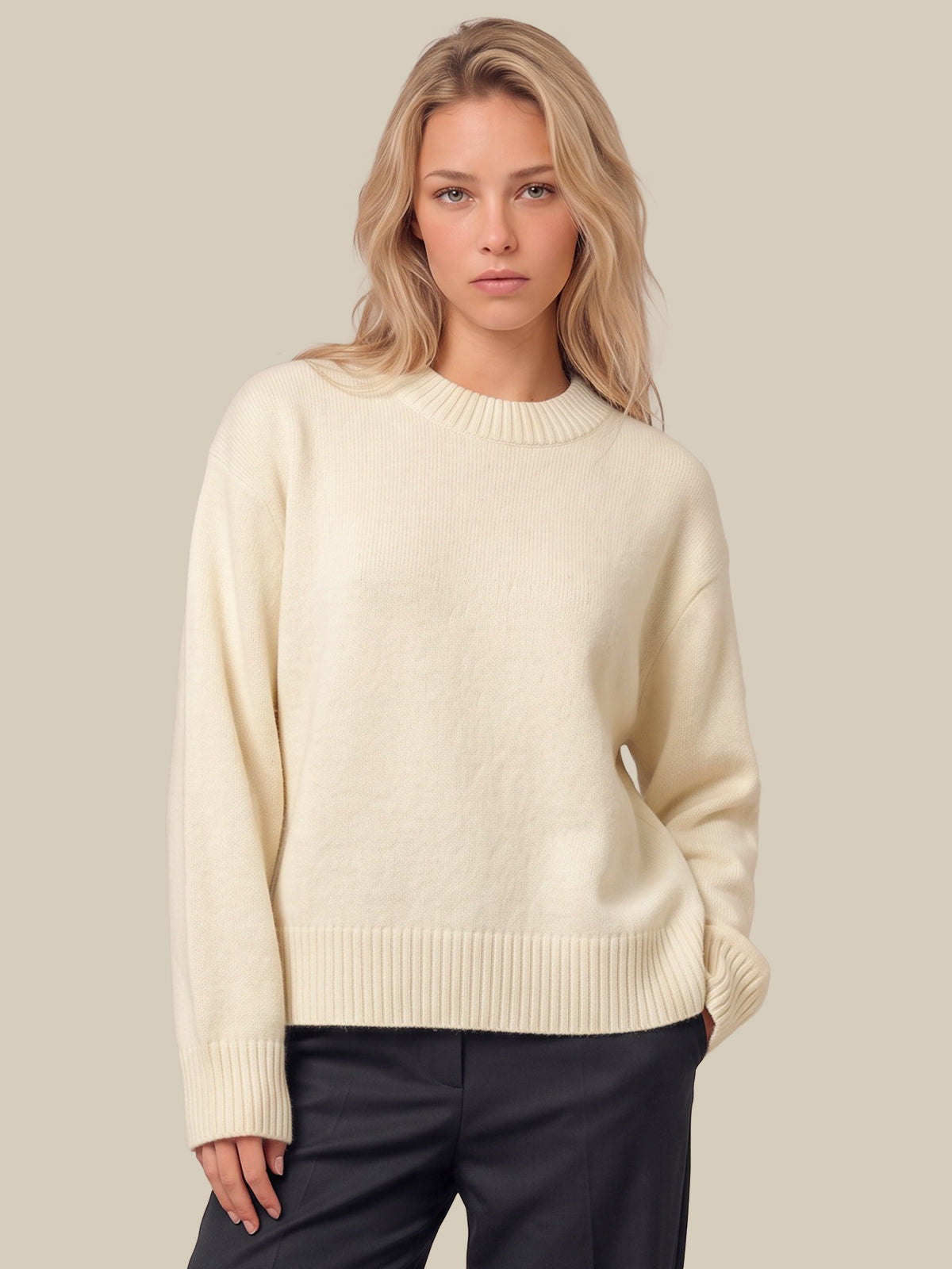 A blonde woman wearing a white-colored knit crewneck sweater and dark charcoal pants, standing against a solid light beige background.