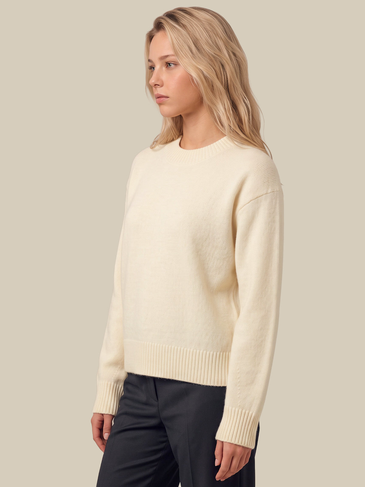 A woman wearing a white-colored knit crewneck sweater and dark trousers, shown from a side profile against a solid light beige background.