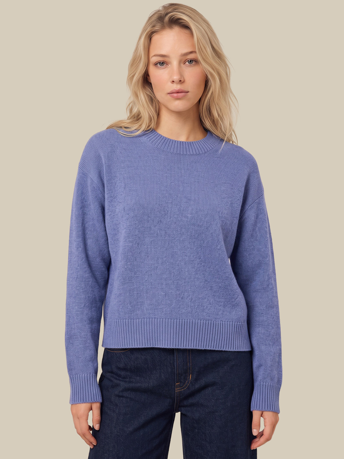 A woman wearing a dusty periwinkle blue crewneck sweater and dark blue jeans, standing against a solid beige background.