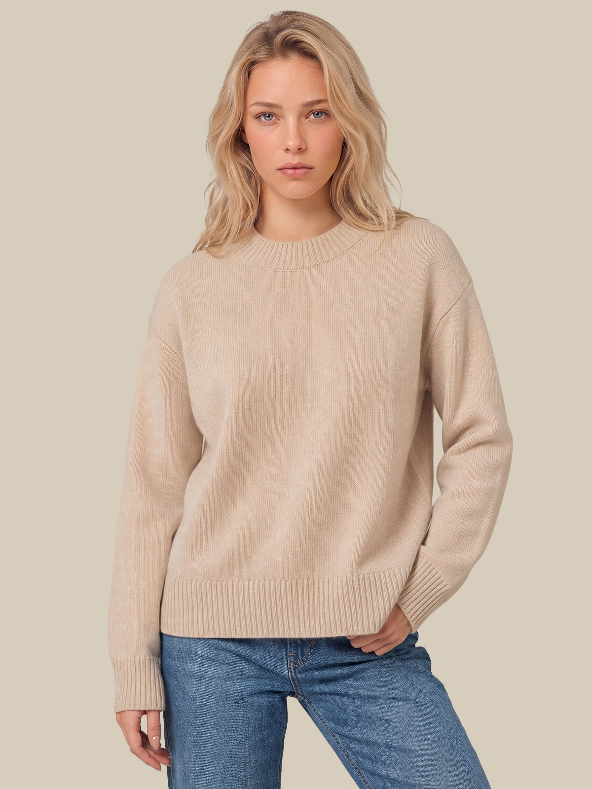 A woman wearing a light beige crewneck knit sweater and blue jeans, standing against a solid light beige background.