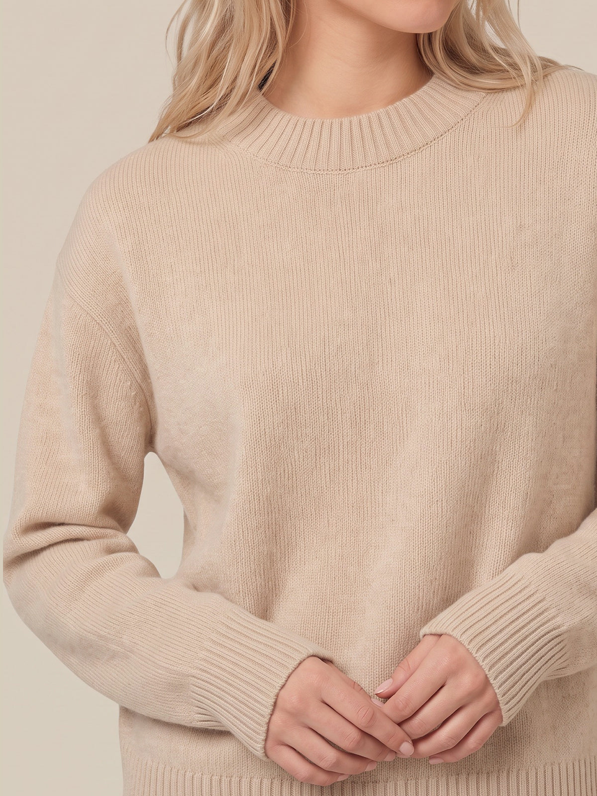 A close-up of a woman wearing a ribbed knit crewneck sweater in a light beige color, with her hands clasped in front.