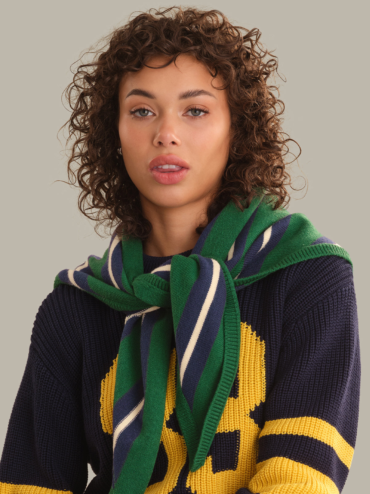 A woman with curly brown hair wearing a navy blue knit sweater with yellow and green graphic details, accessorized with a green, navy, and cream striped knit scarf, against a solid light gray background.