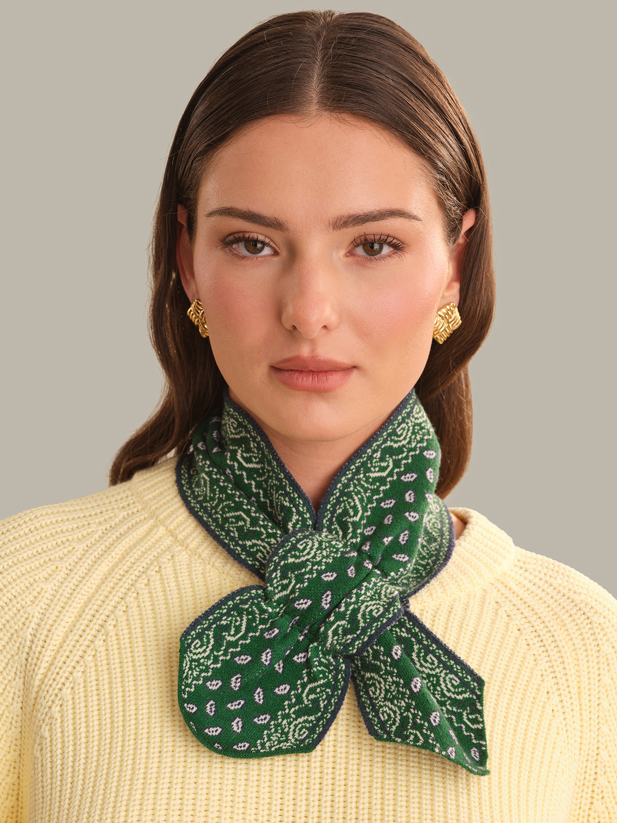 A woman wearing a green and white paisley patterned silk scarf tied around her neck, paired with a cream-colored ribbed knit sweater and gold earrings, against a solid gray background.