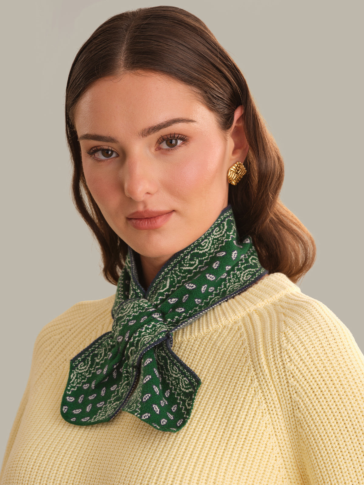 A woman wearing a green paisley bandana-style neck scarf tied in a bow around her collar, paired with a cream-colored ribbed knit sweater against a solid light gray background.