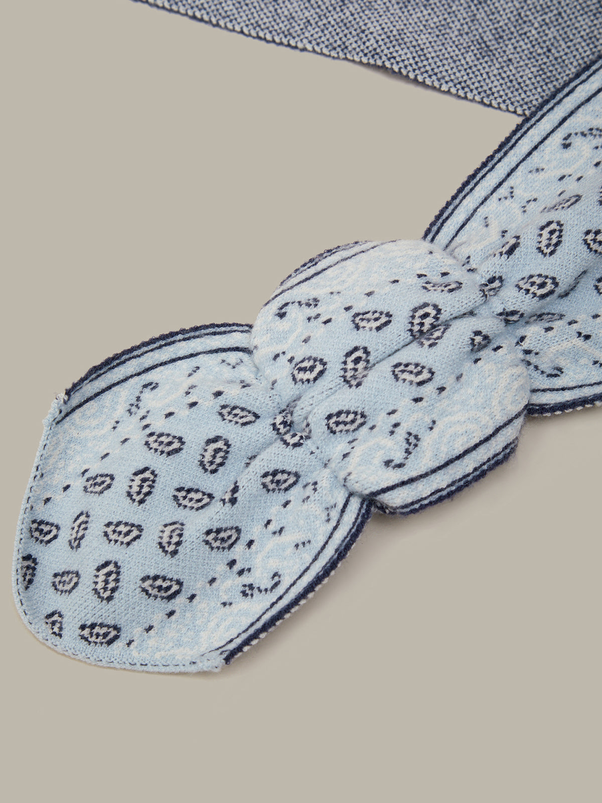 A light blue paisley bandana tie with dark navy borders, laid flat on a neutral beige background.