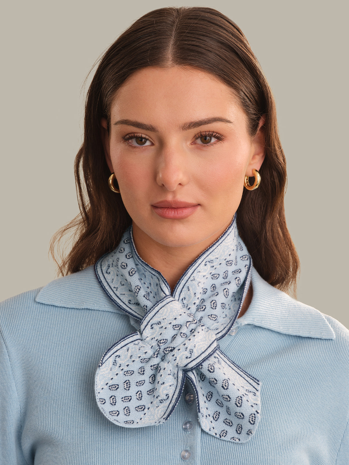 A woman wearing a light blue ribbed knit top and a light blue patterned scarf tied around her neck, with gold hoop earrings, against a solid beige background.