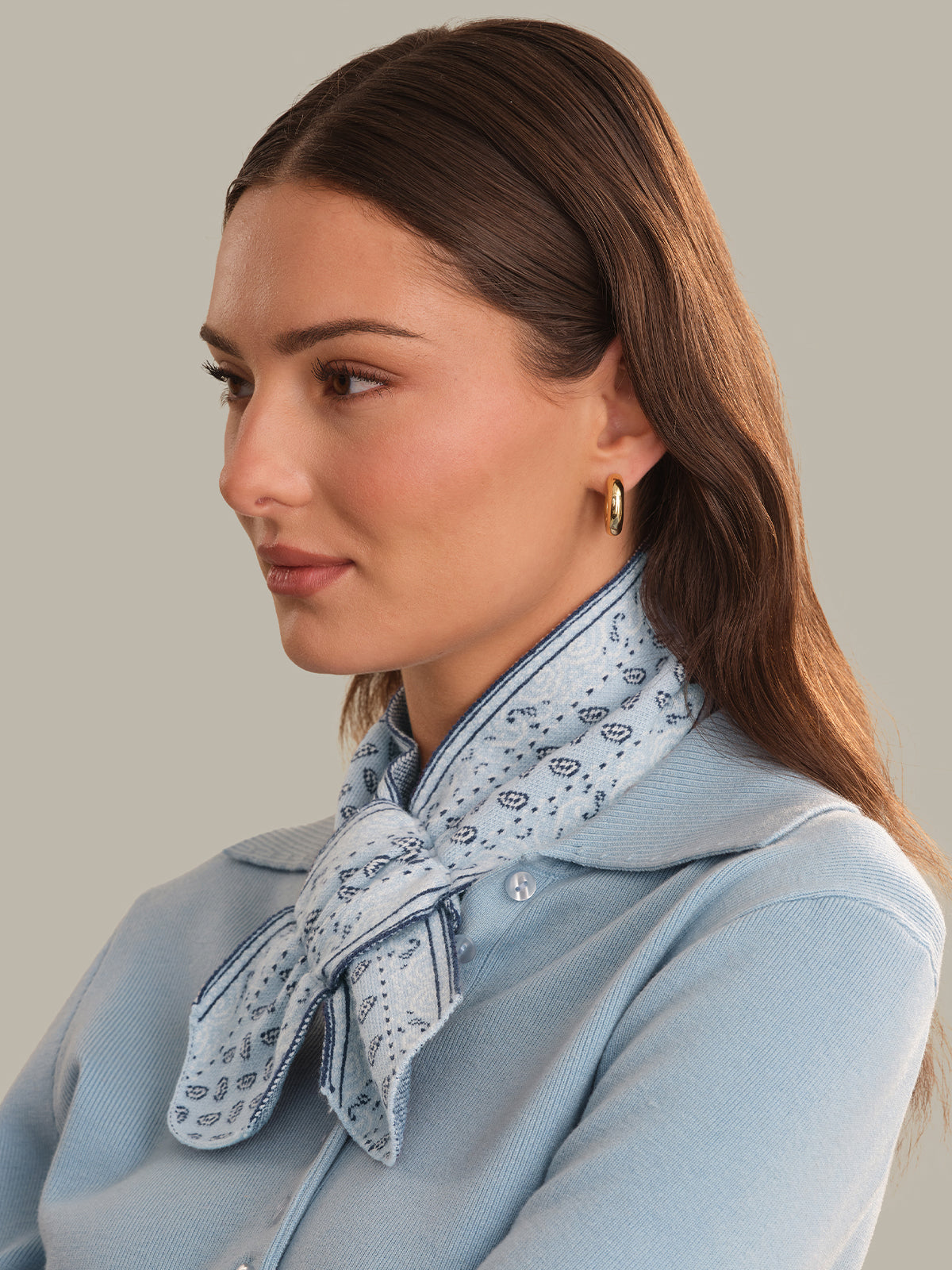 A woman in profile wearing a light blue knit cardigan and a light blue paisley scarf tied around her neck, against a solid warm gray background.