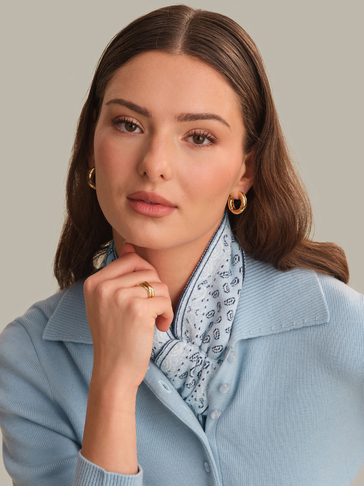 A woman with brown hair wearing a light blue ribbed cardigan, a light blue and white paisley scarf, and gold hoop earrings, posing against a solid beige background.