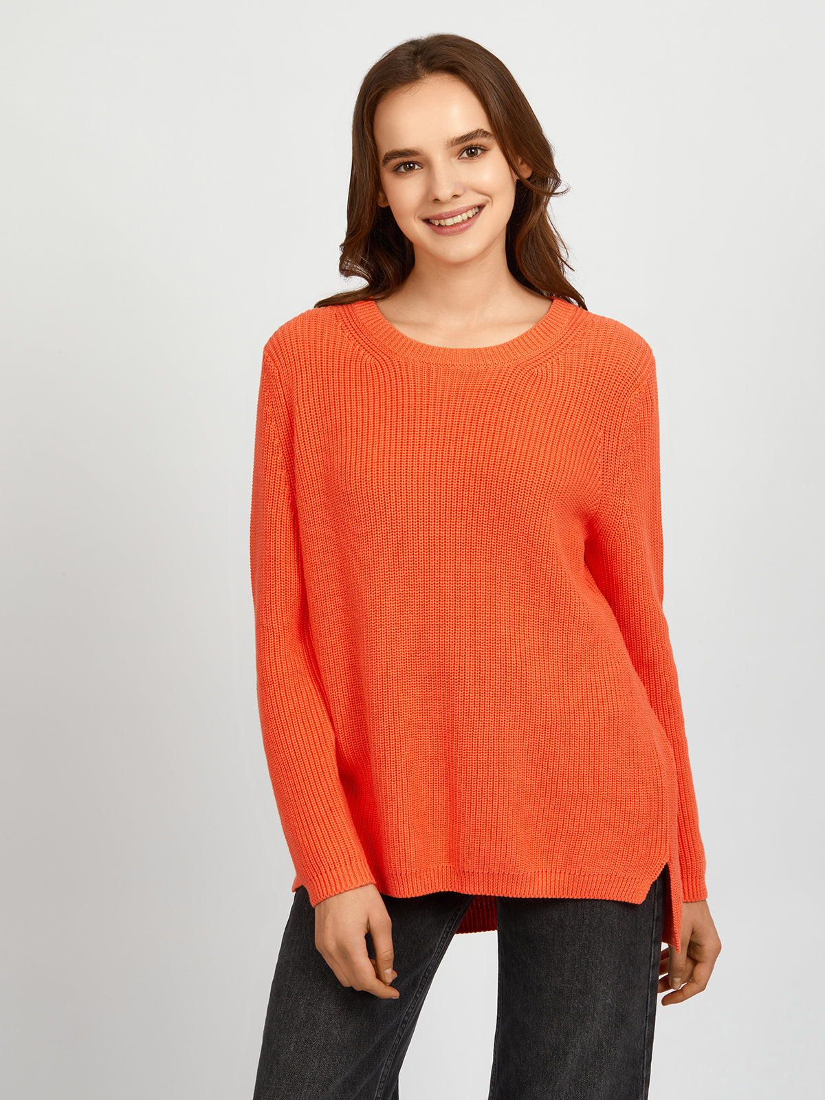 A person stands against a plain light gray background, wearing a relaxed fit, long-sleeve, bright orange 100% cotton shaker stitch sweater by 525 America and dark pants, with a slight smile and relaxed posture.