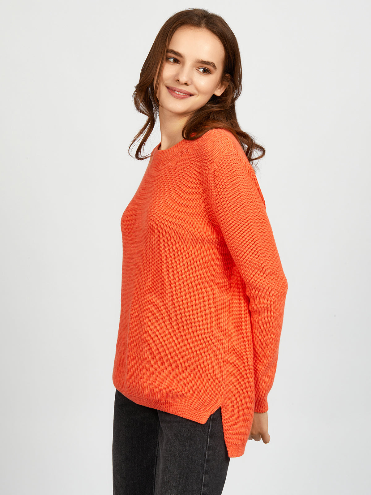 A person with long brown hair wears a 525 America orange 100% cotton shaker stitch knit sweater and dark pants, standing against a plain light gray background, looking slightly to the side and smiling.