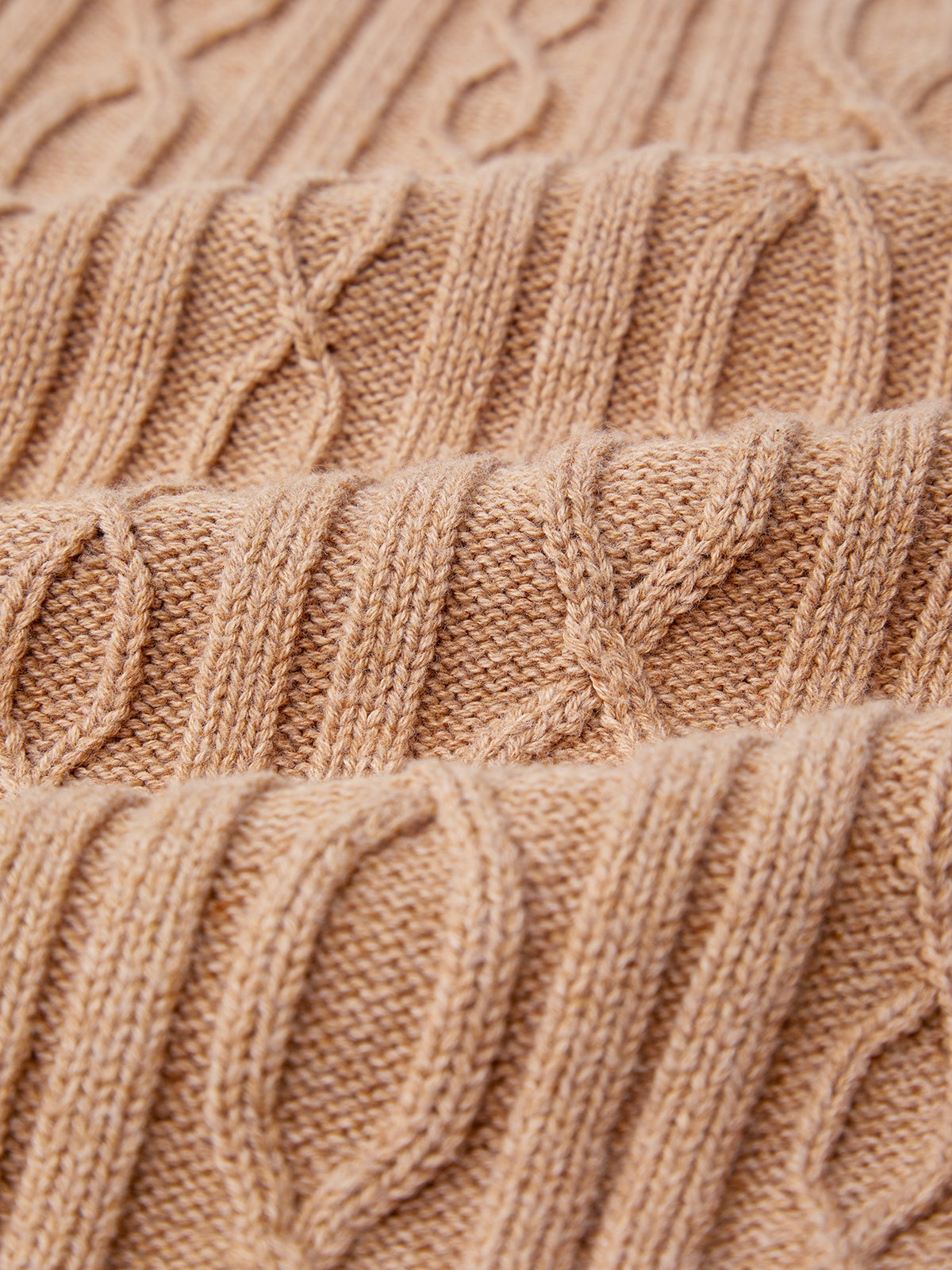 Close-up of a beige cable knit sweater by 525 America, showcasing a repeating pattern. Made from 100% cotton, the texture features intertwining yarn, forming raised, rounded designs running parallel across the fabrics surface.