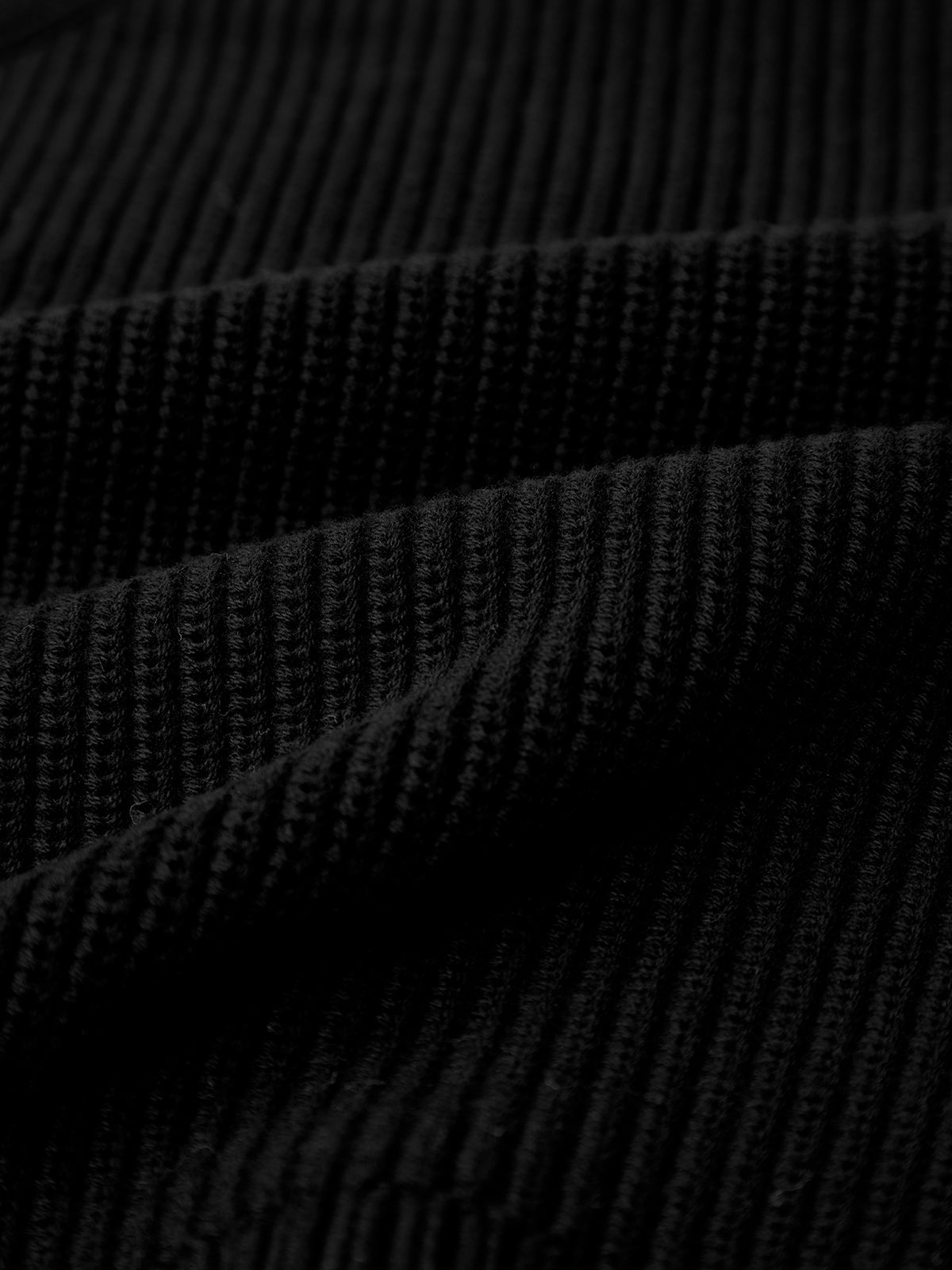 Close-up of black ribbed fabric on a 525 America cotton shaker stitch sweater with horizontal folds. The texture displays raised lines, forming subtle ridges. The lighting highlights its structure, enhanced by a fitted silhouette and raglan sleeves.
