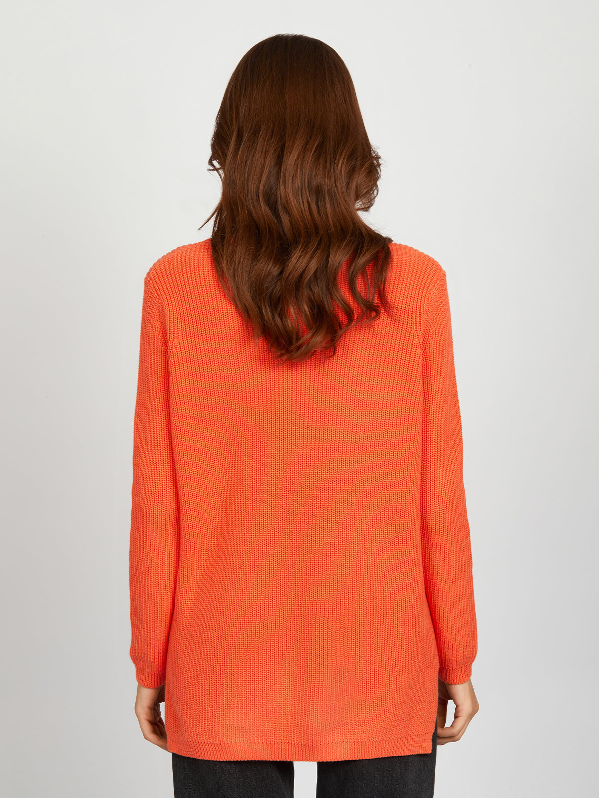 With their back to the camera, a person with long wavy brown hair models a bright orange 525 America shaker-stitch knit sweater and dark pants against a plain light background.