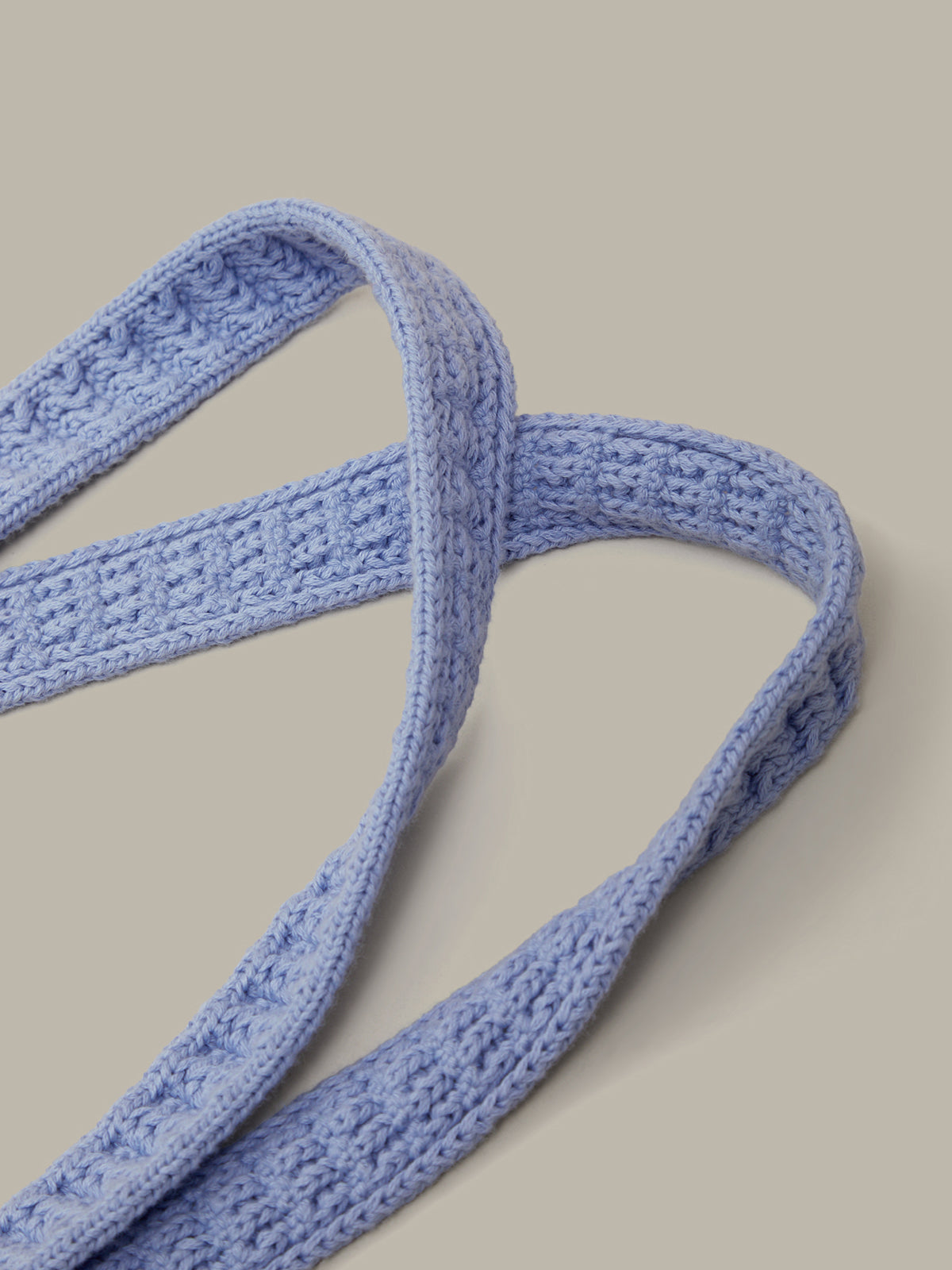 A close-up of a light blue knitted strap with a textured cable stitch pattern, arranged in loops against a solid beige background.
