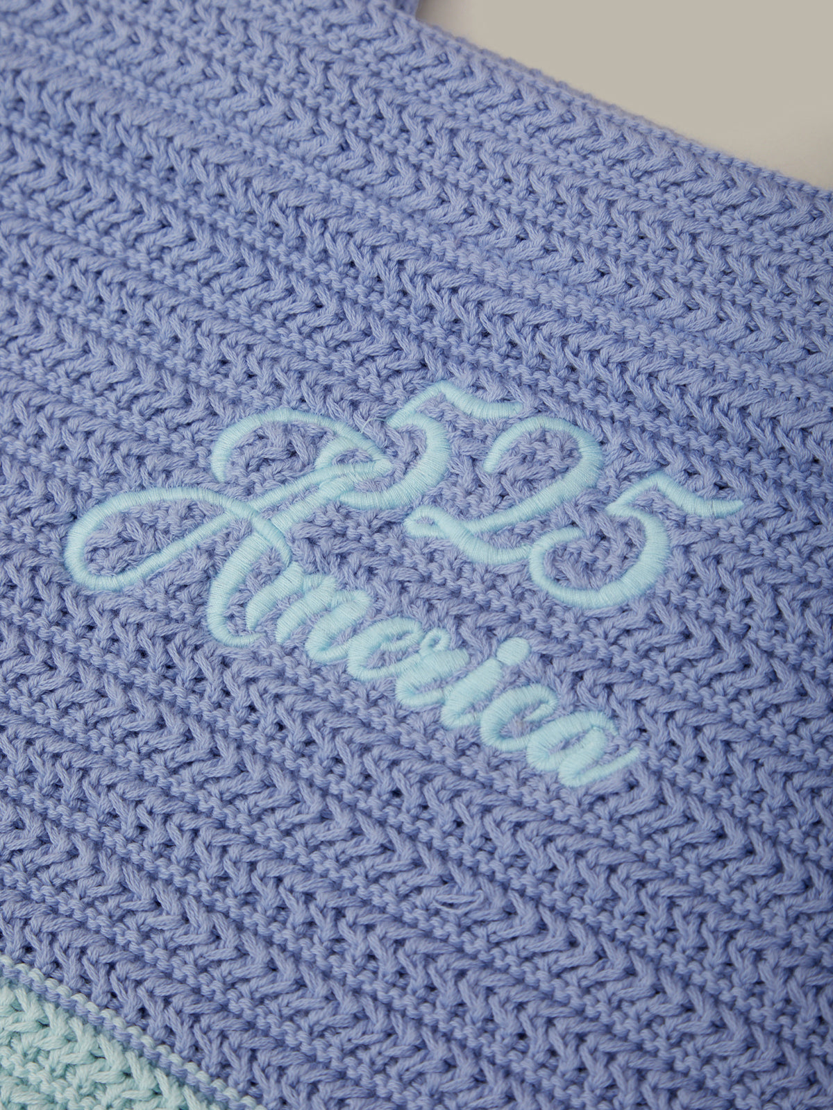 A close-up of a blue crocheted textile with light blue embroidered script reading '525 America' across the center.