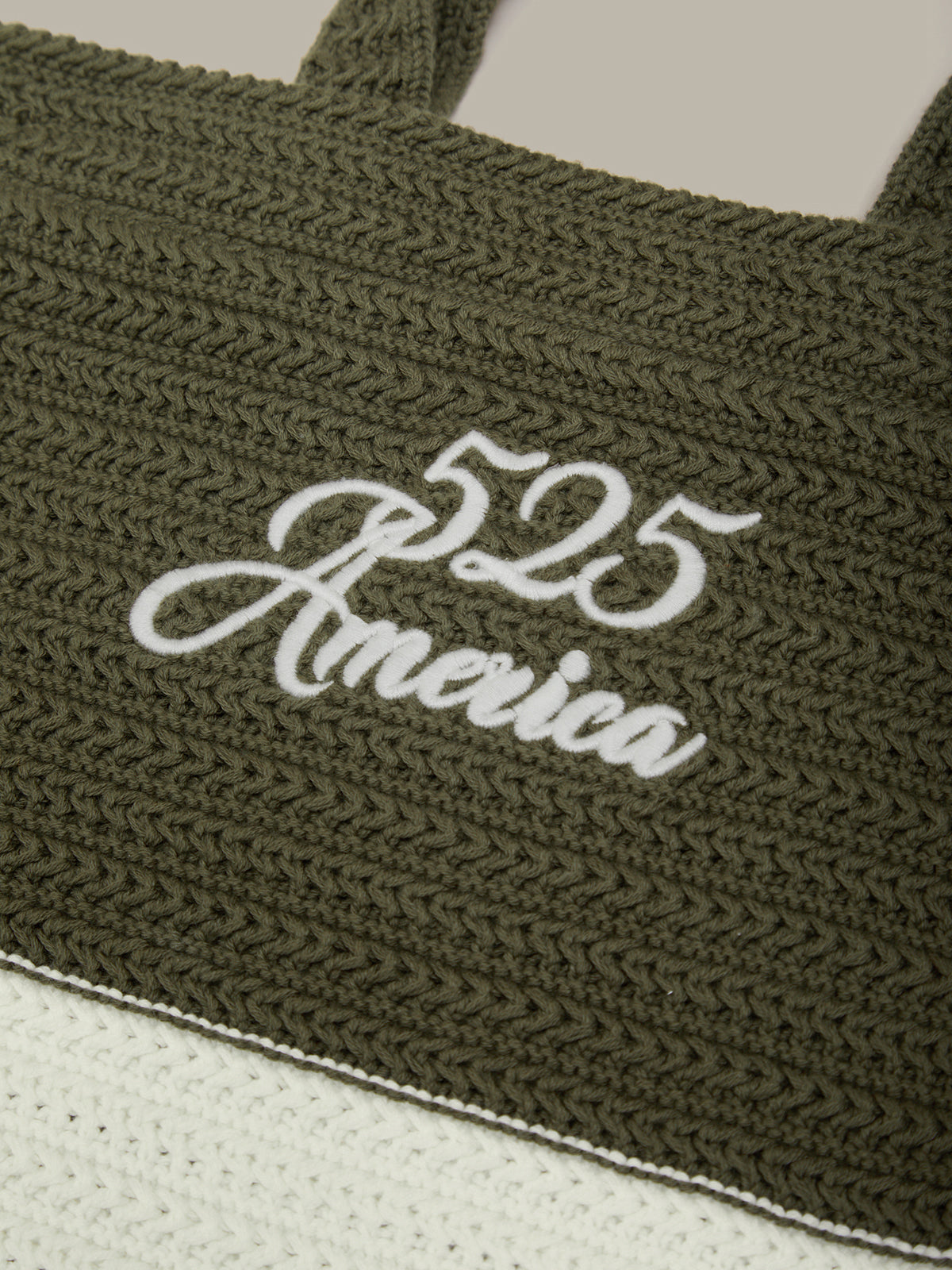 Close-up of an olive green woven tote bag with white embroidered text reading '525 America' on a light neutral background.