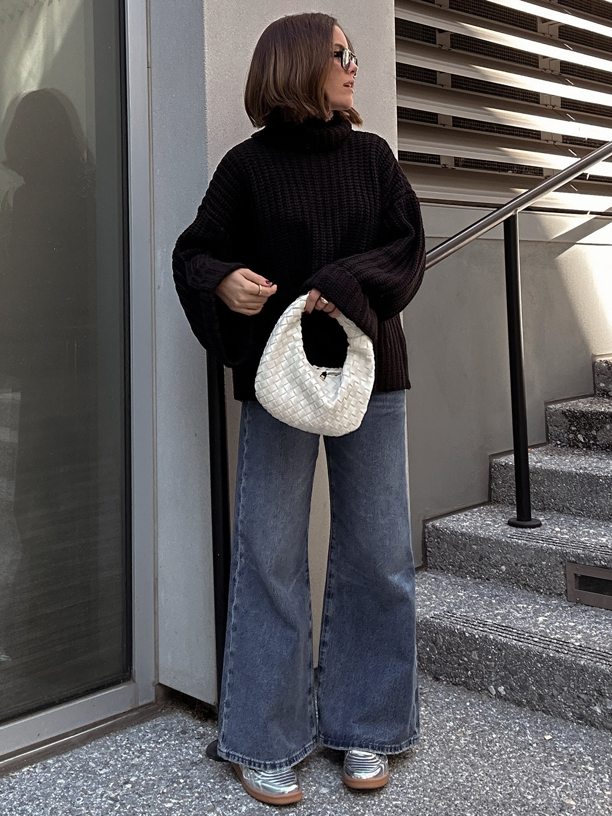 A person stands on concrete steps, wearing a cozy 525 America turtleneck with oversized sleeves, wide-leg jeans, and silver sneakers. They hold a white woven handbag and wear sunglasses in front of a modern glass and metal structure.