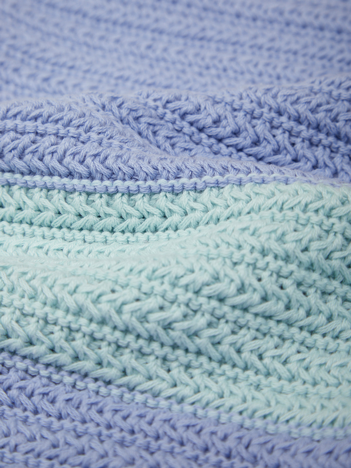 A close-up of a knitted blanket or textile with horizontal stripes in soft lavender, dusty blue, mint green, and pale blue, showing detailed knit texture.
