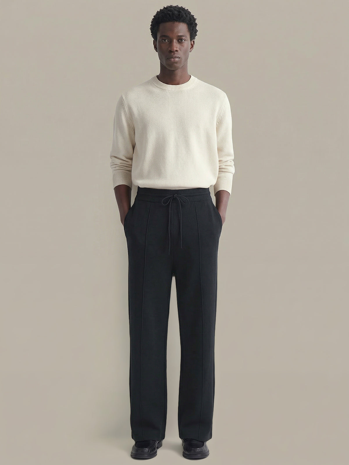 Man wearing a cream sweater and black pants against a beige background