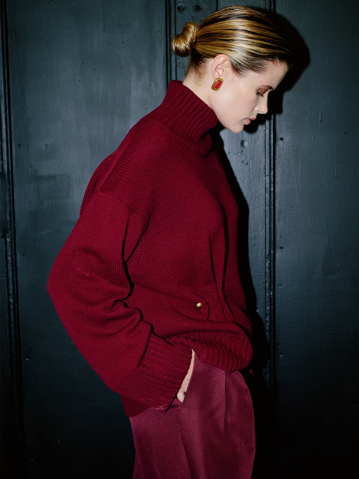 A woman in a dark studio setting wearing an oversized ribbed turtleneck sweater in deep burgundy with matching trousers and red geometric earrings, posed against a dark paneled background.