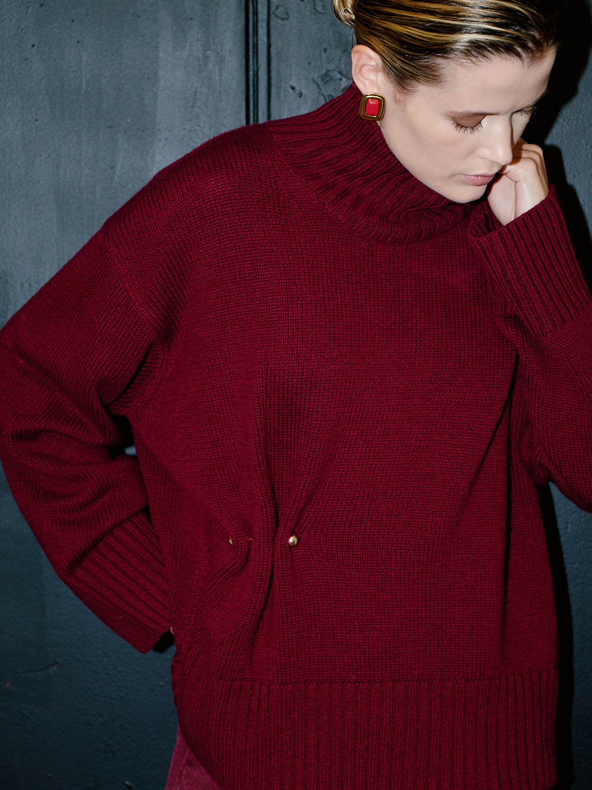 A woman wearing a chunky knit turtleneck sweater in deep burgundy red, featuring small gold button details at the waist, posed against a dark textured wall.
