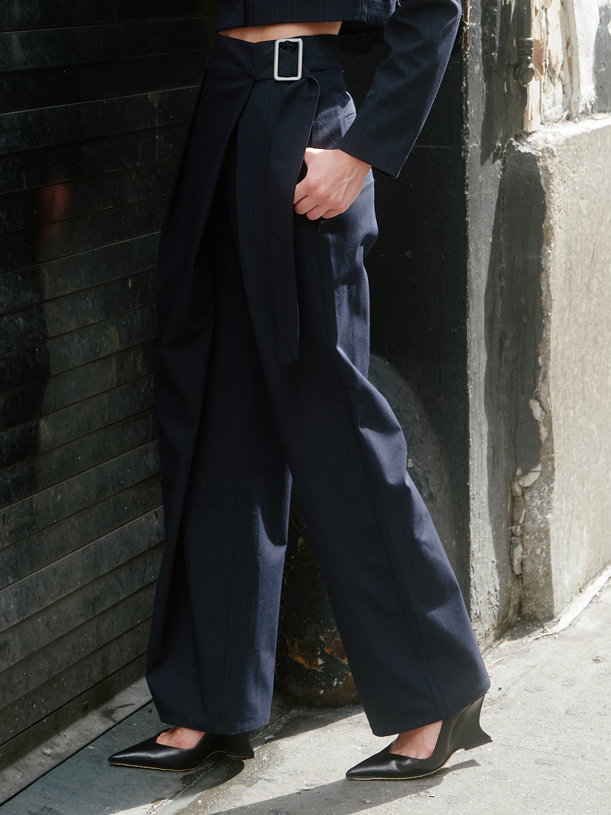 A person wearing dark navy wide-leg trousers with a wrap-style front and a silver rectangular belt buckle, paired with black pointed-toe heels, standing against a textured dark wall.