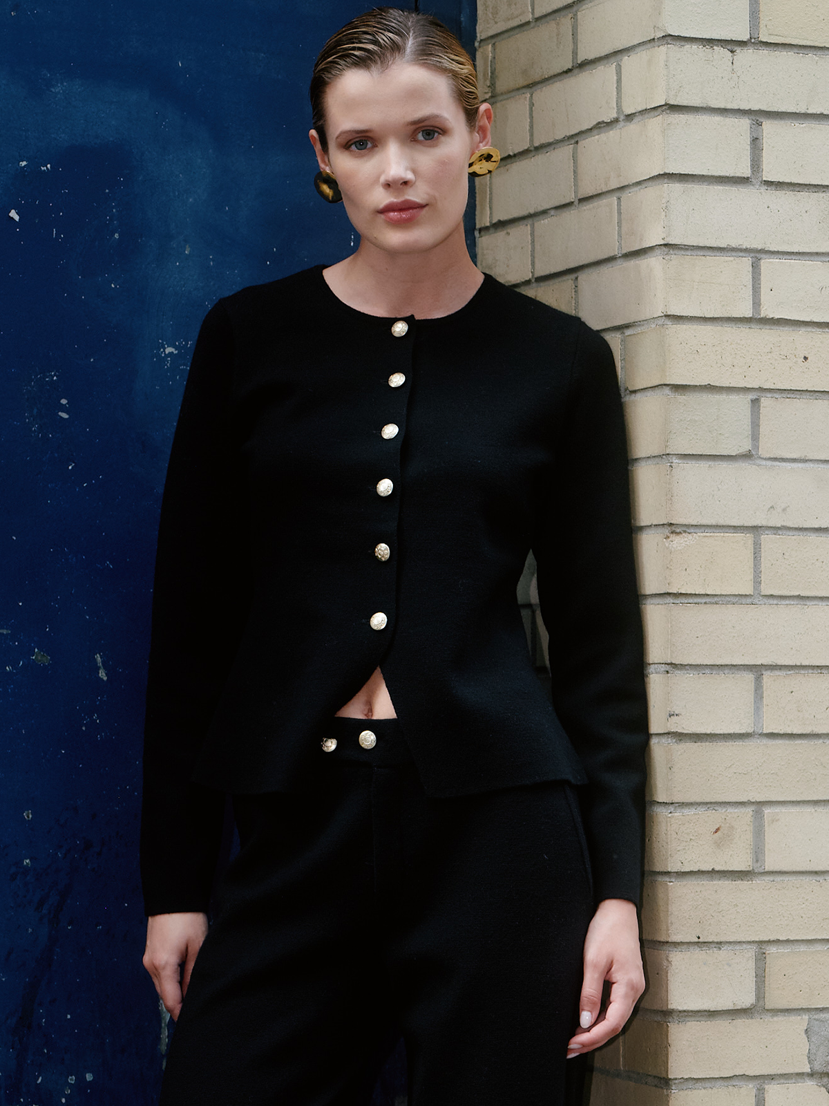 A model wearing a black cropped jacket with gold buttons and matching black trousers, standing against a split blue and beige brick wall.