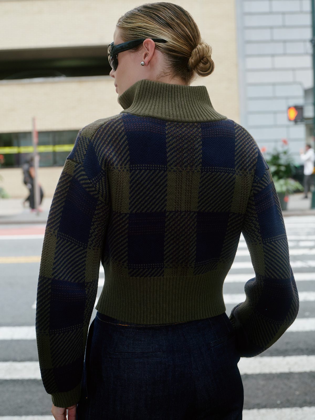 A woman viewed from behind wearing a cropped knit sweater with a large check pattern in olive green and navy blue, paired with dark jeans and sunglasses, standing on a city crosswalk.