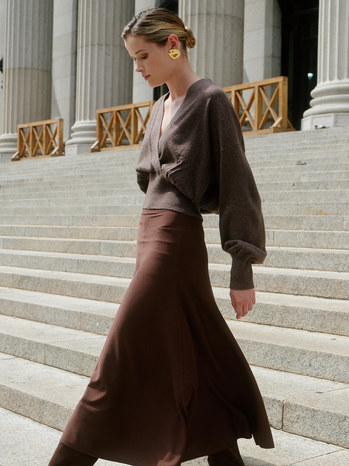 A woman walks down grand stone steps in front of classical columns, wearing a brown knit wrap-style sweater and a matching brown maxi skirt. She is also wearing prominent gold earrings.