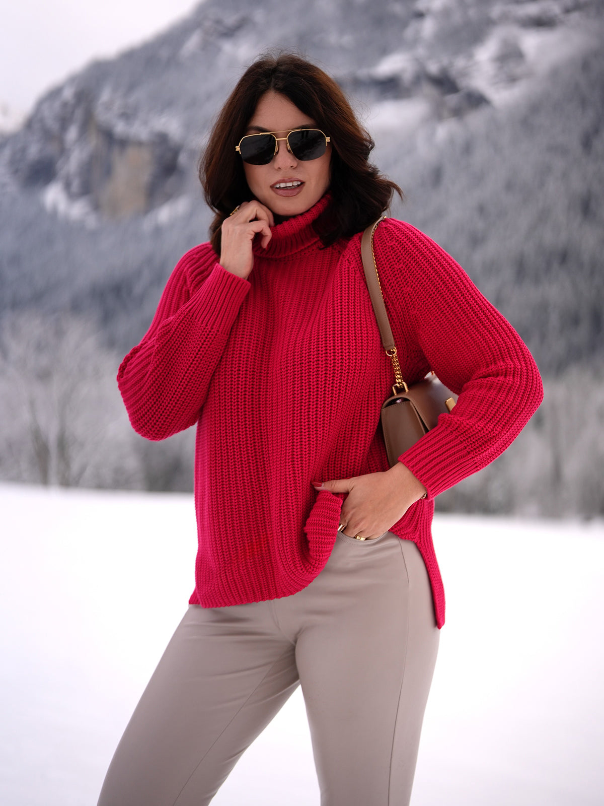 In a snowy landscape, someone exudes cozy vibes in a bright pink 525 America 100% cotton turtleneck sweater and beige pants. Sporting sunglasses and carrying a brown shoulder bag, theyre framed by snow-covered trees and majestic mountains.