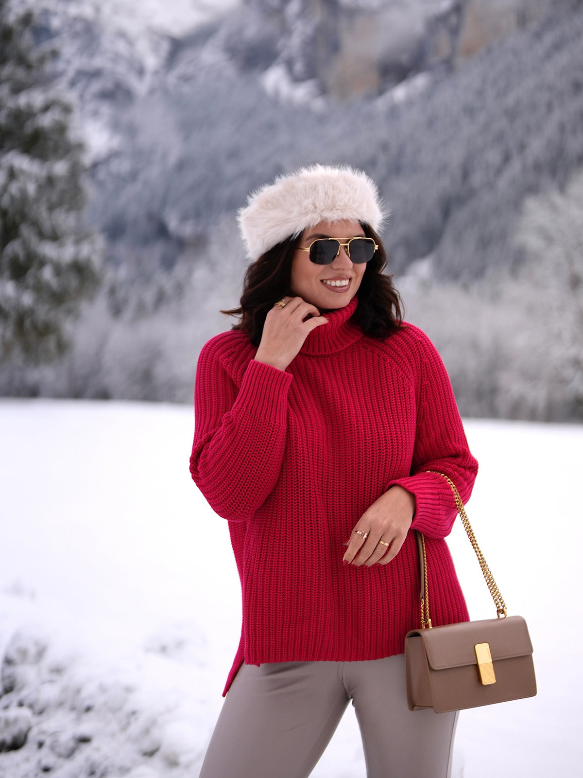 Dressed in a cozy red knit sweater from 525 America, sunglasses, a fur hat, and carrying a brown handbag, the person stands amid a snowy landscape with mountains and trees in the background.
