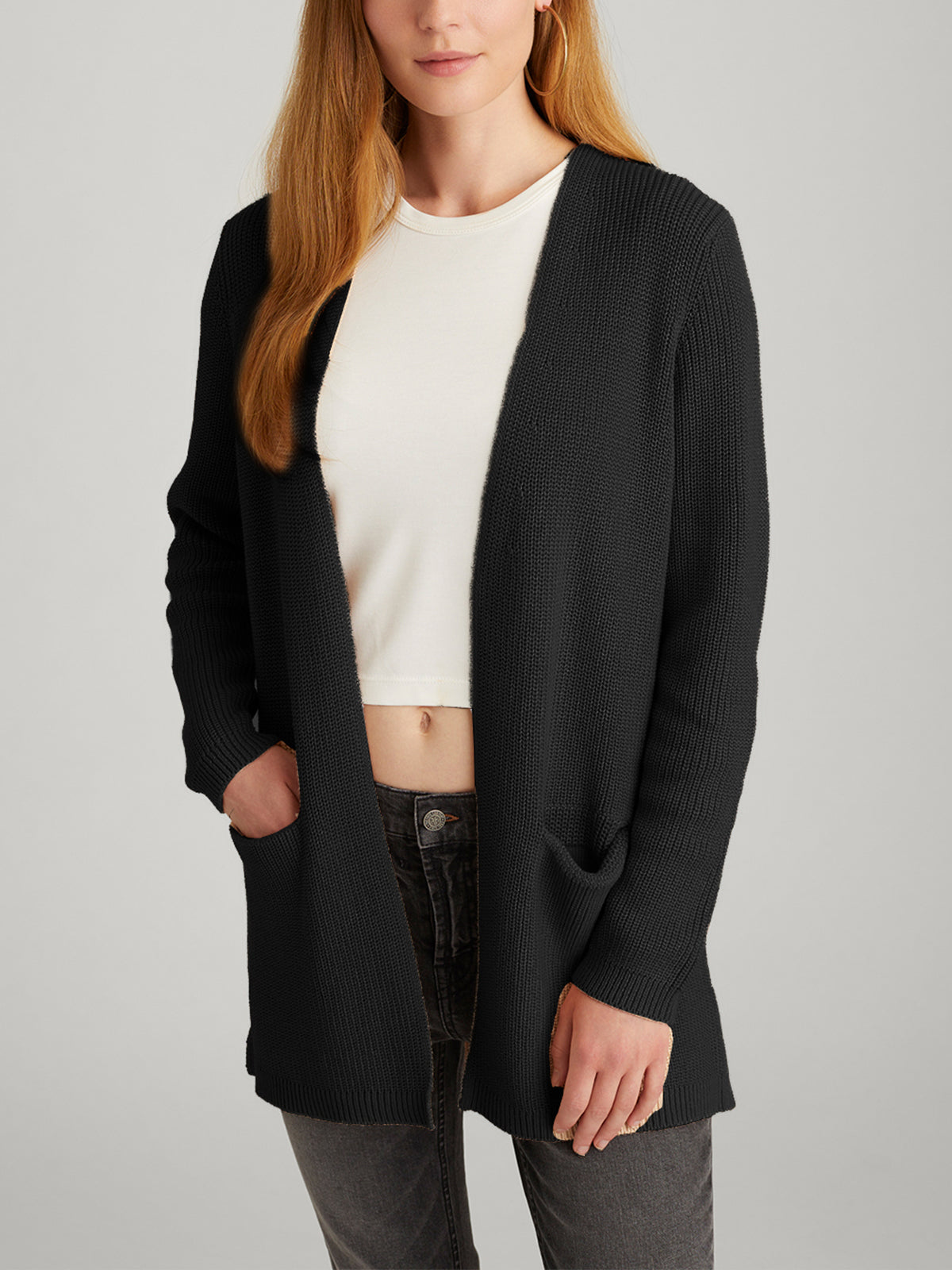 A person in an Emma: Shaker Stitch Open Cardigan, featuring pockets, is layered over a white crop top with dark jeans. The black cardigan complements their long, light brown hair against a plain background.