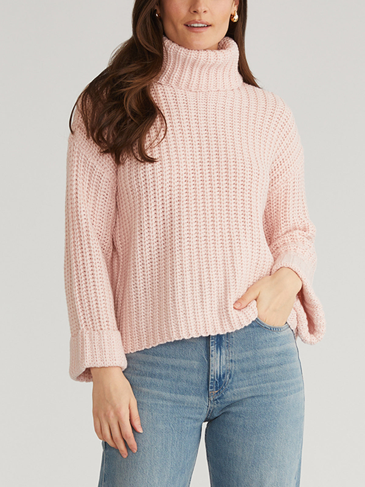 A person wearing 525 Americas light pink Turtleneck Shaker Pullover with textured pattern and oversized sleeves pairs it effortlessly with blue jeans. The cozy turtleneck accentuates their long brown hair as one hand rests casually in a pocket against the plain background.