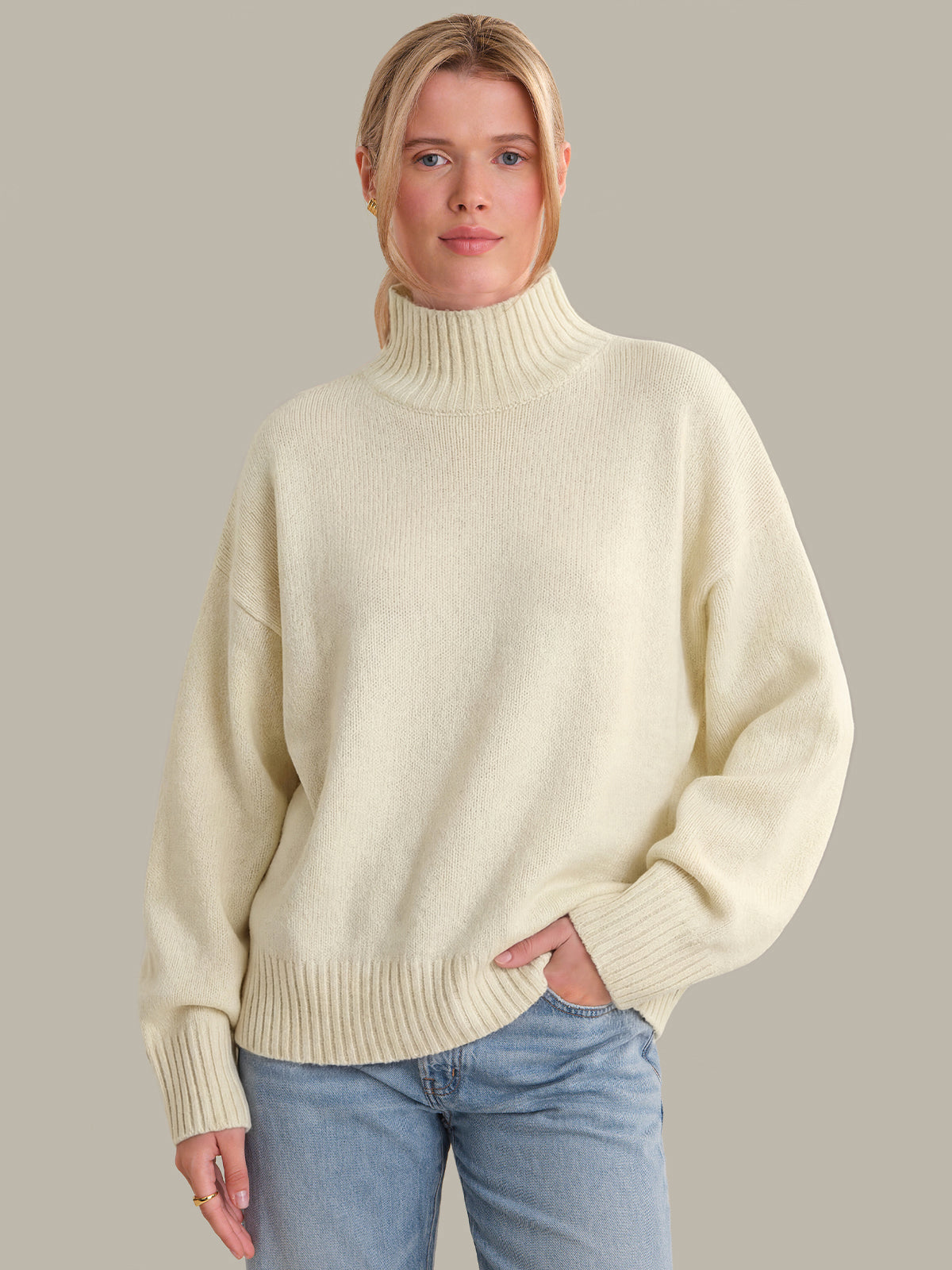 Woman wearing a cream turtleneck sweater and blue jeans against a neutral background