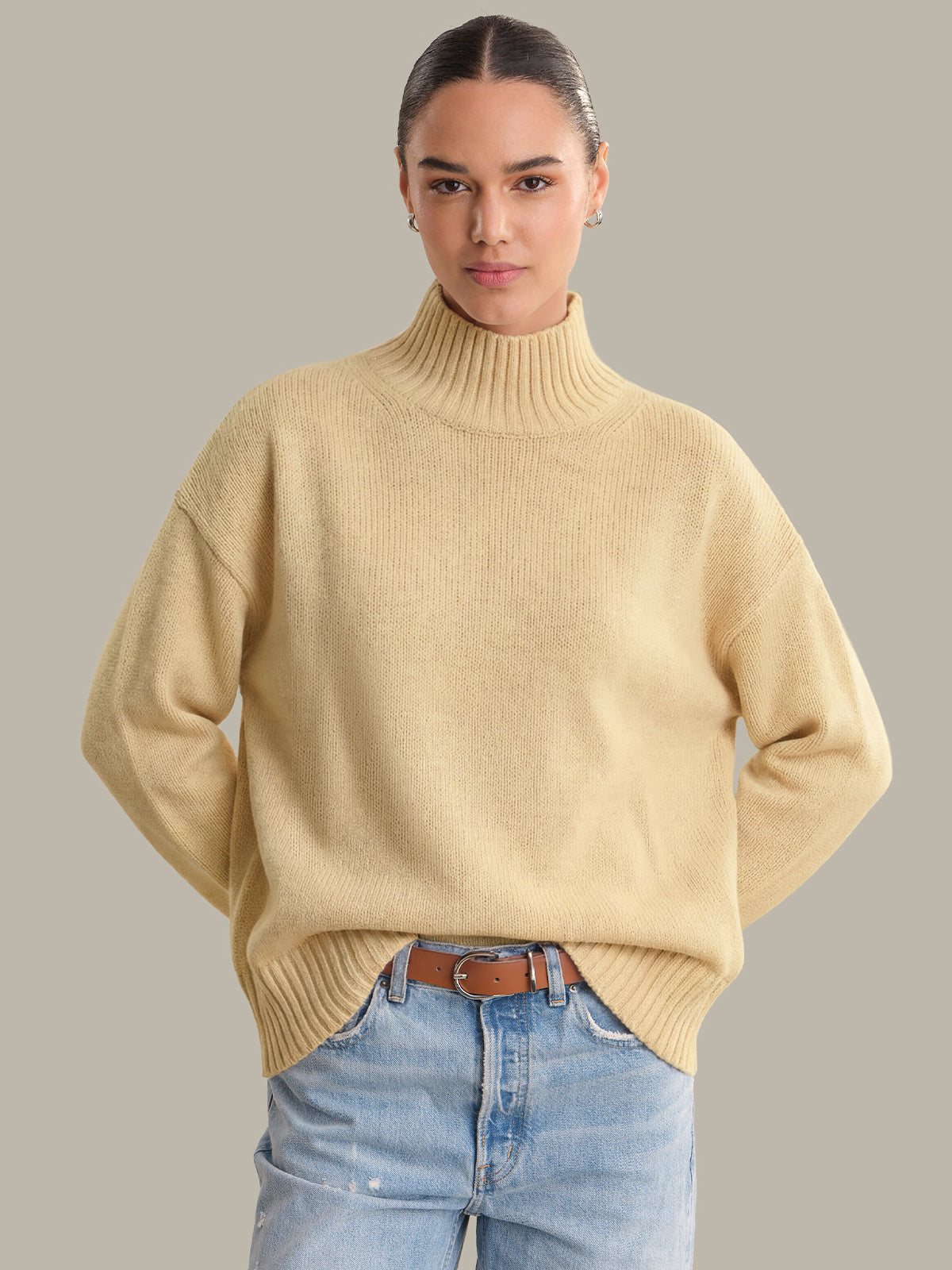 Woman wearing a beige sweater and blue jeans against a plain background