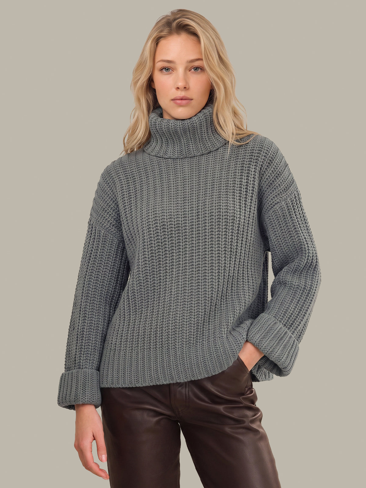 A woman wearing a chunky gray ribbed turtleneck sweater and dark brown pants, standing against a solid light gray background.