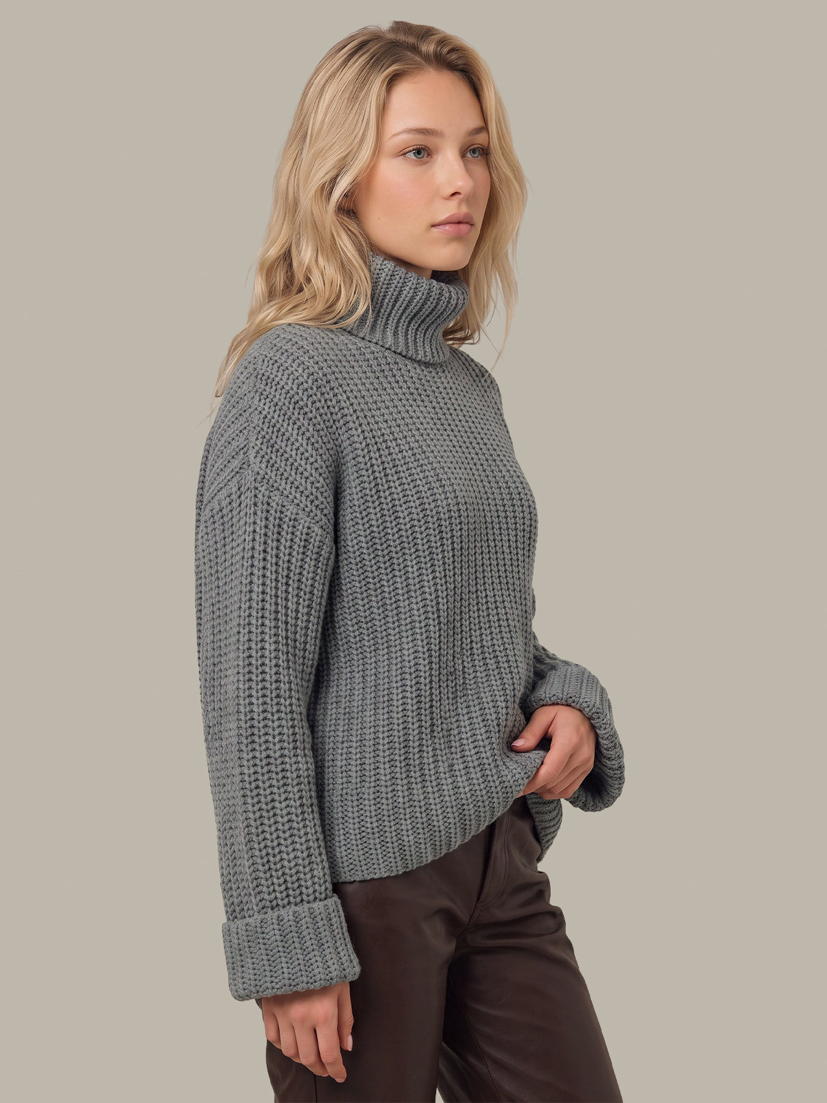 A woman wearing a chunky gray ribbed turtleneck sweater and dark brown pants, shown in a side profile against a solid light gray background.