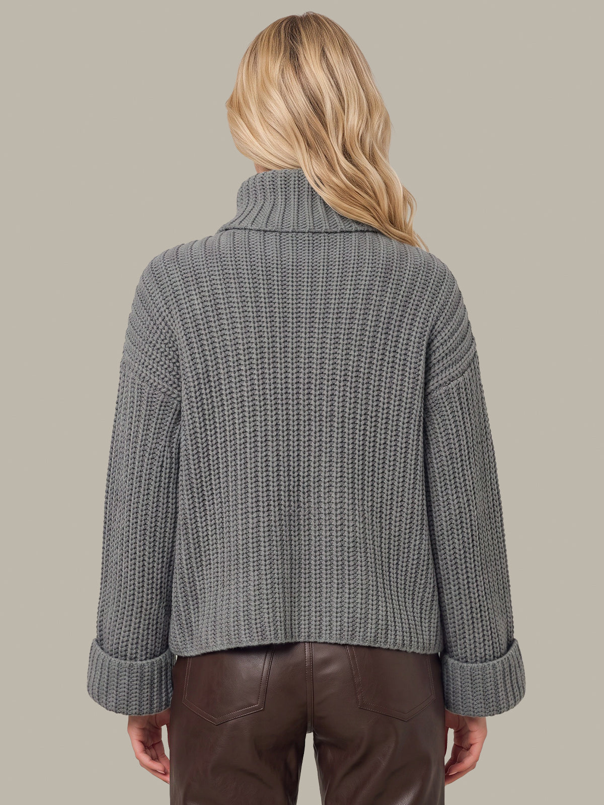 A woman viewed from the back wearing a gray ribbed knit turtleneck sweater and brown pants against a solid taupe background.