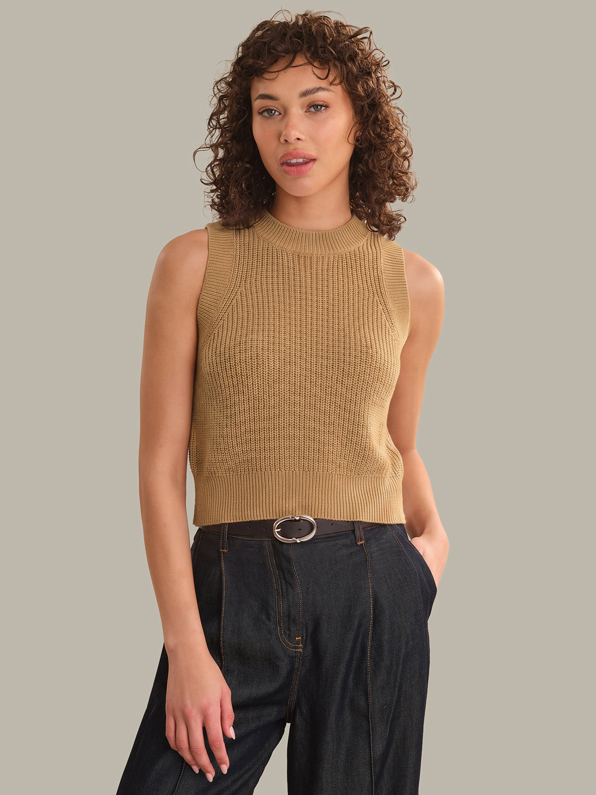 Woman wearing a beige sleeveless knitted top with black pants on a plain background