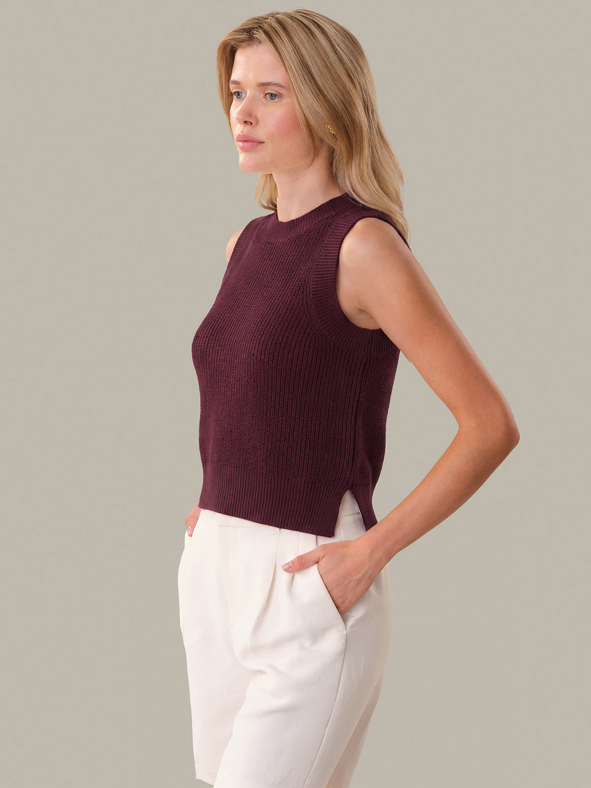 Woman wearing a sleeveless burgundy top and white pants against a neutral background