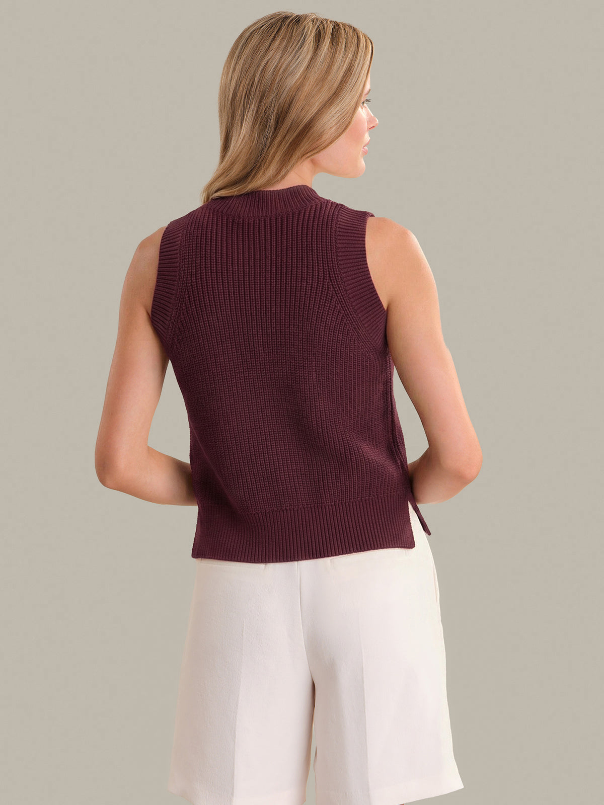 Woman wearing a sleeveless burgundy sweater with white pants on a neutral background