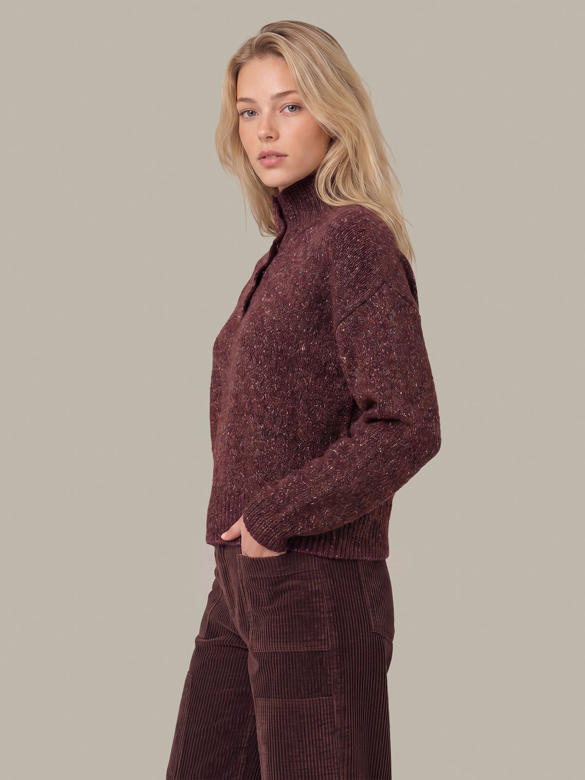 A blonde woman wearing a textured, heathered burgundy knit mock-neck sweater and matching corduroy pants, shown in a side profile against a solid taupe background.