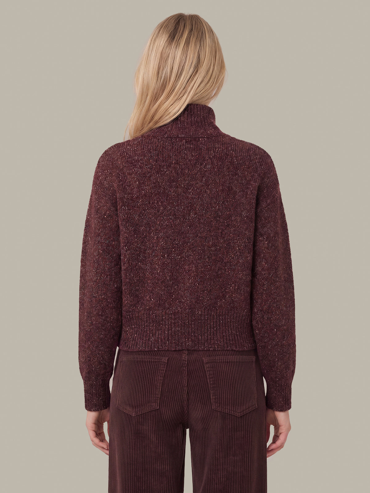A woman shown from the back wearing a textured, heathered dark burgundy turtleneck sweater and matching corduroy trousers against a solid taupe background.