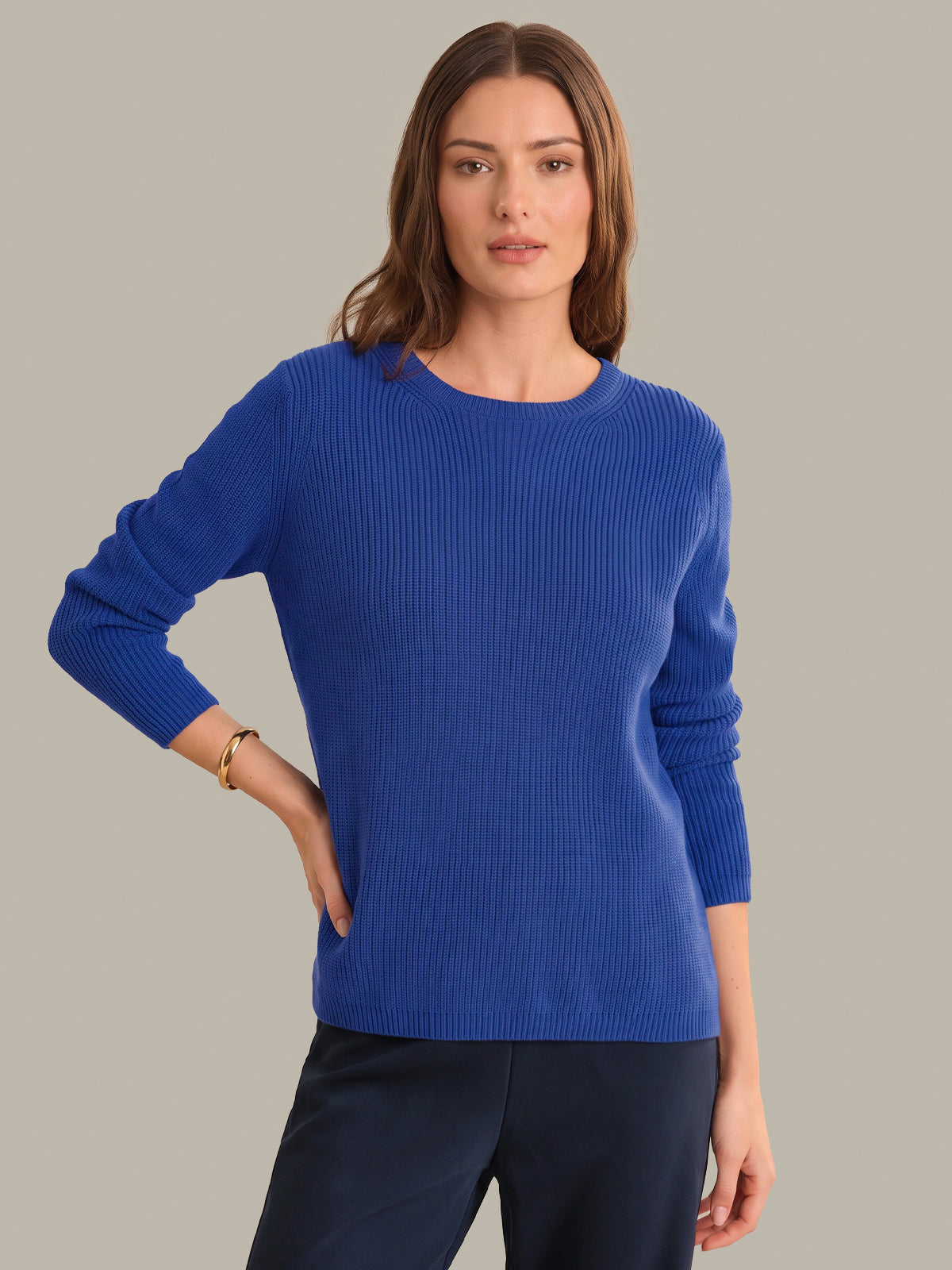 A woman modeling a royal blue ribbed knit sweater with three-quarter sleeves, paired with dark navy trousers, against a solid light gray background.